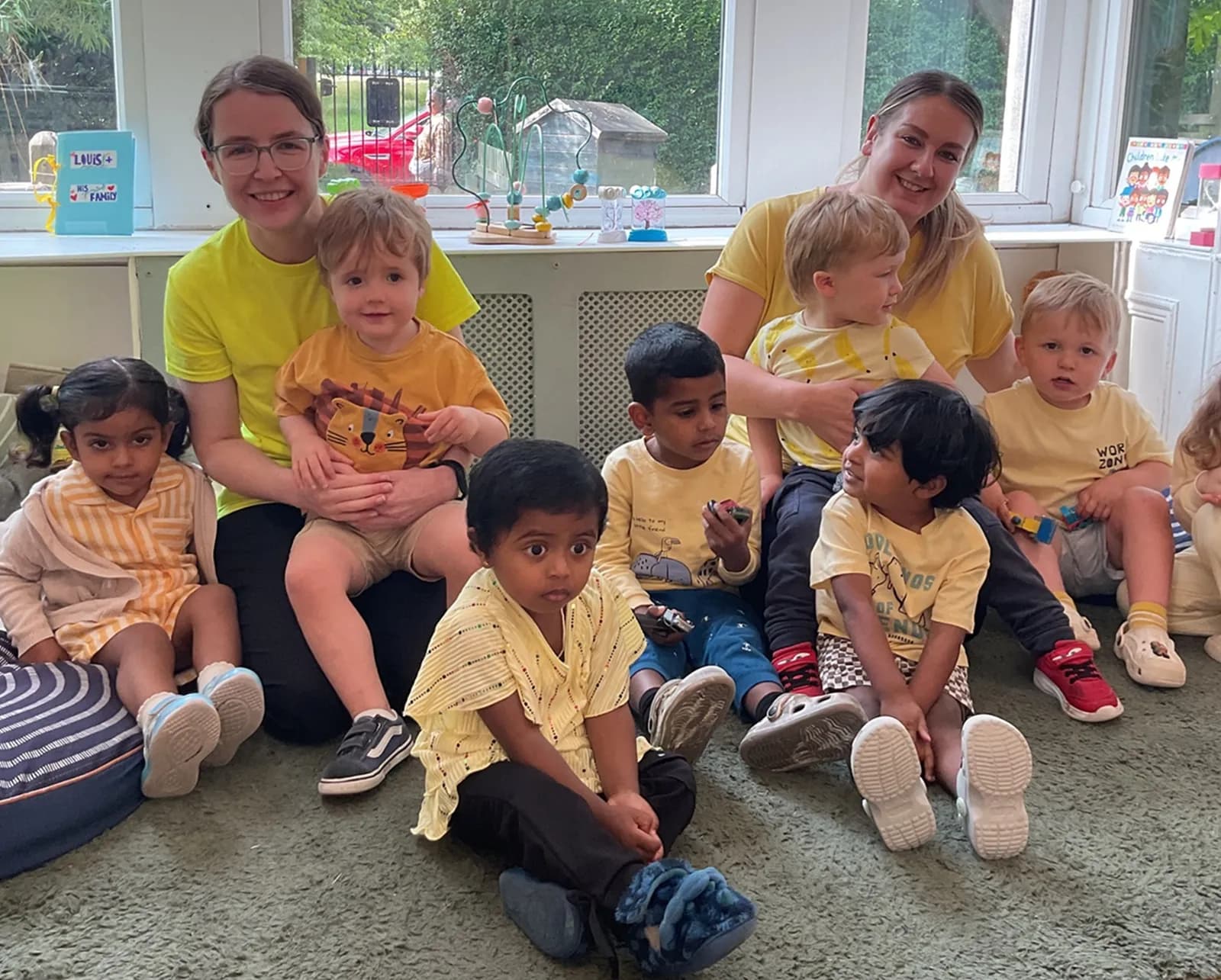 Two women wearing yellow shirts with a group of young children sitting on a rug and a blue cushion in front of a window, all smiling and looking forward.